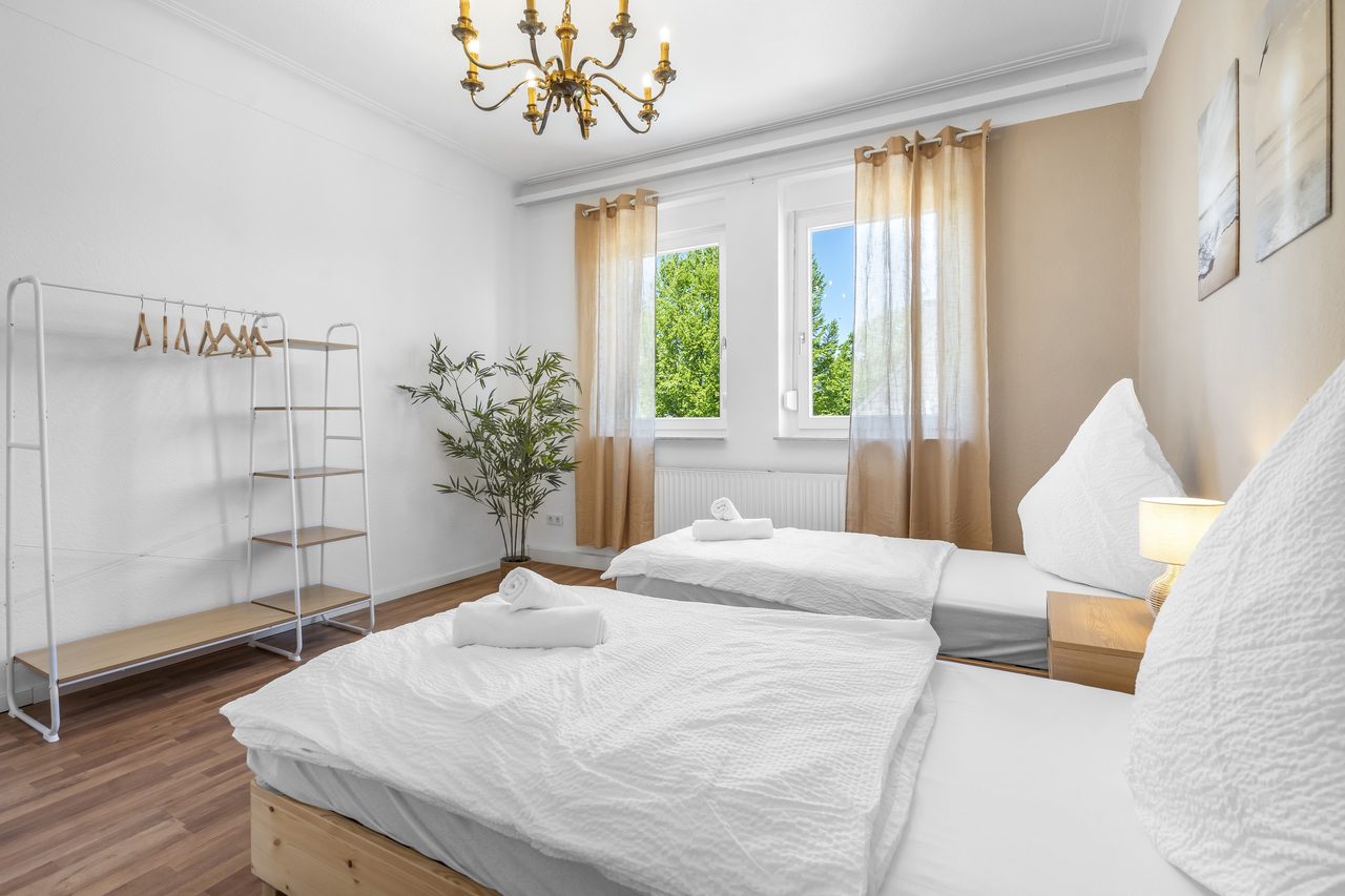 Helles Schlafzimmer mit zwei weiß bezogenen Betten, Messing-Kronleuchter, Kleiderständer und einer grünen Zimmerpflanze am sonnigen Fenster.