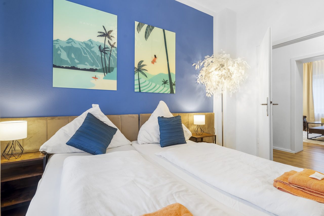 Breites Doppelbett mit weißer Bettwäsche und blauen Kissen vor einer blauen Wand mit zwei Palmenbildern, beide Nachttischlampen brennen.