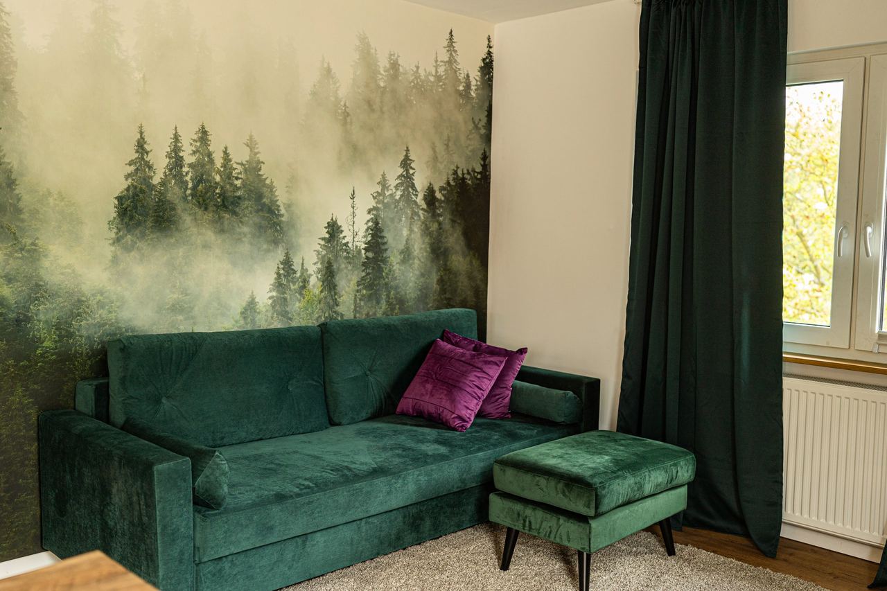 Grünes Samtsofa mit Hocker vor einer Fototapete mit Nebelwald, rechts ein Fenster mit gelbem Herbstlaub dahinter.