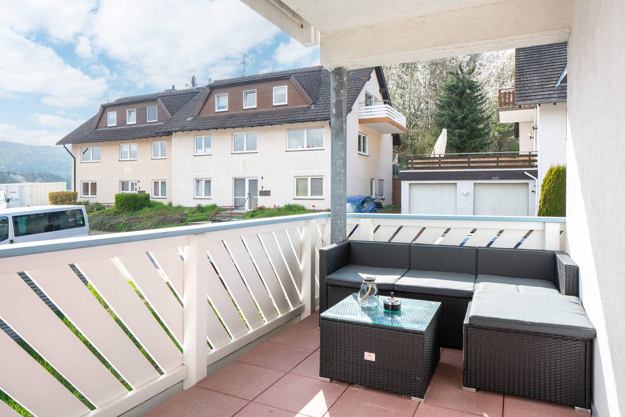 Überdachter Balkon mit Rattan-Ecksofa und Glastisch, dahinter Nachbarhäuser, Nadelbäume und bewaldete Hügel.