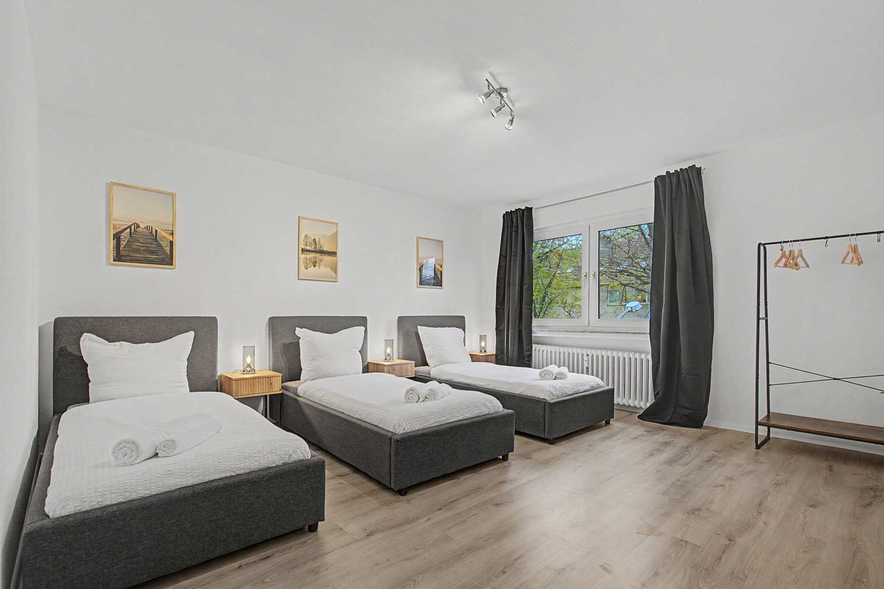 Schlafzimmer mit drei grauen Einzelbetten, Nachttischen mit Lampen, Fenster mit dunklen Vorhängen und Kleiderständer