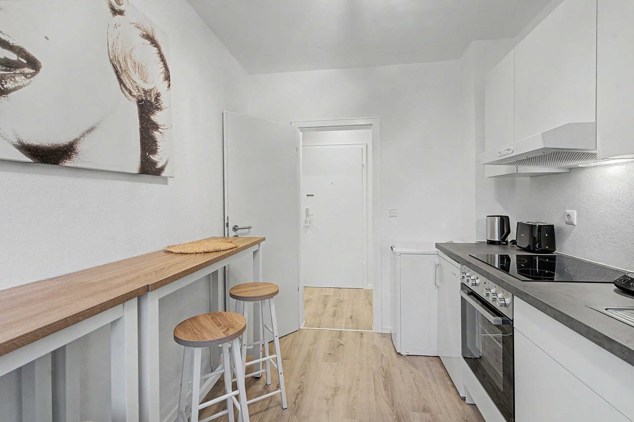 Schmale helle Küche mit einer langen Holztheke an der linken Wand und zwei Barhockern, rechts eine weiße Küchenzeile mit Ceranfeld, Backofen, Wasserkocher und Toaster, hinten der Blick durch den Flur zur Wohnungstür.