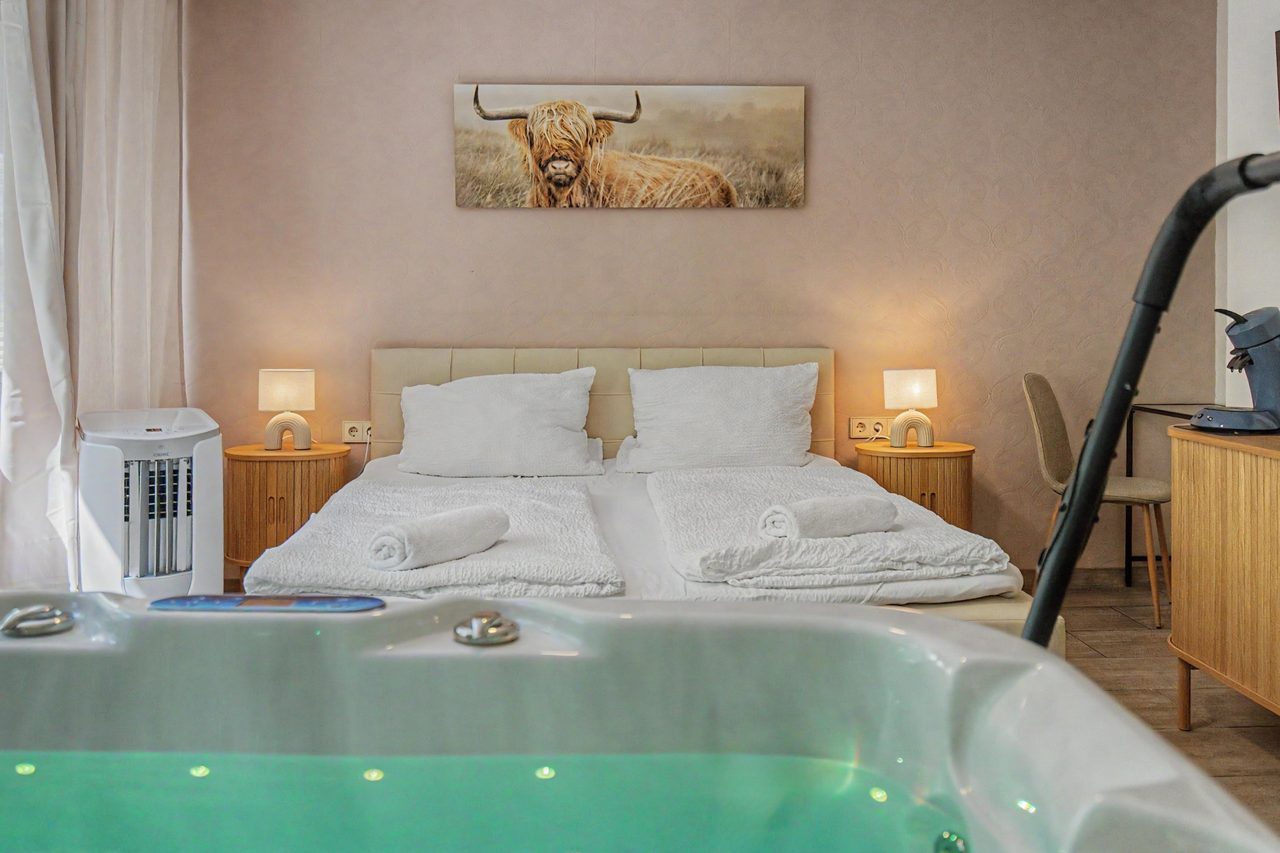 Doppelbett mit weißer Bettwäsche und zwei brennenden Nachttischlampen, im Vordergrund der türkis beleuchtete Whirlpool im selben Zimmer.