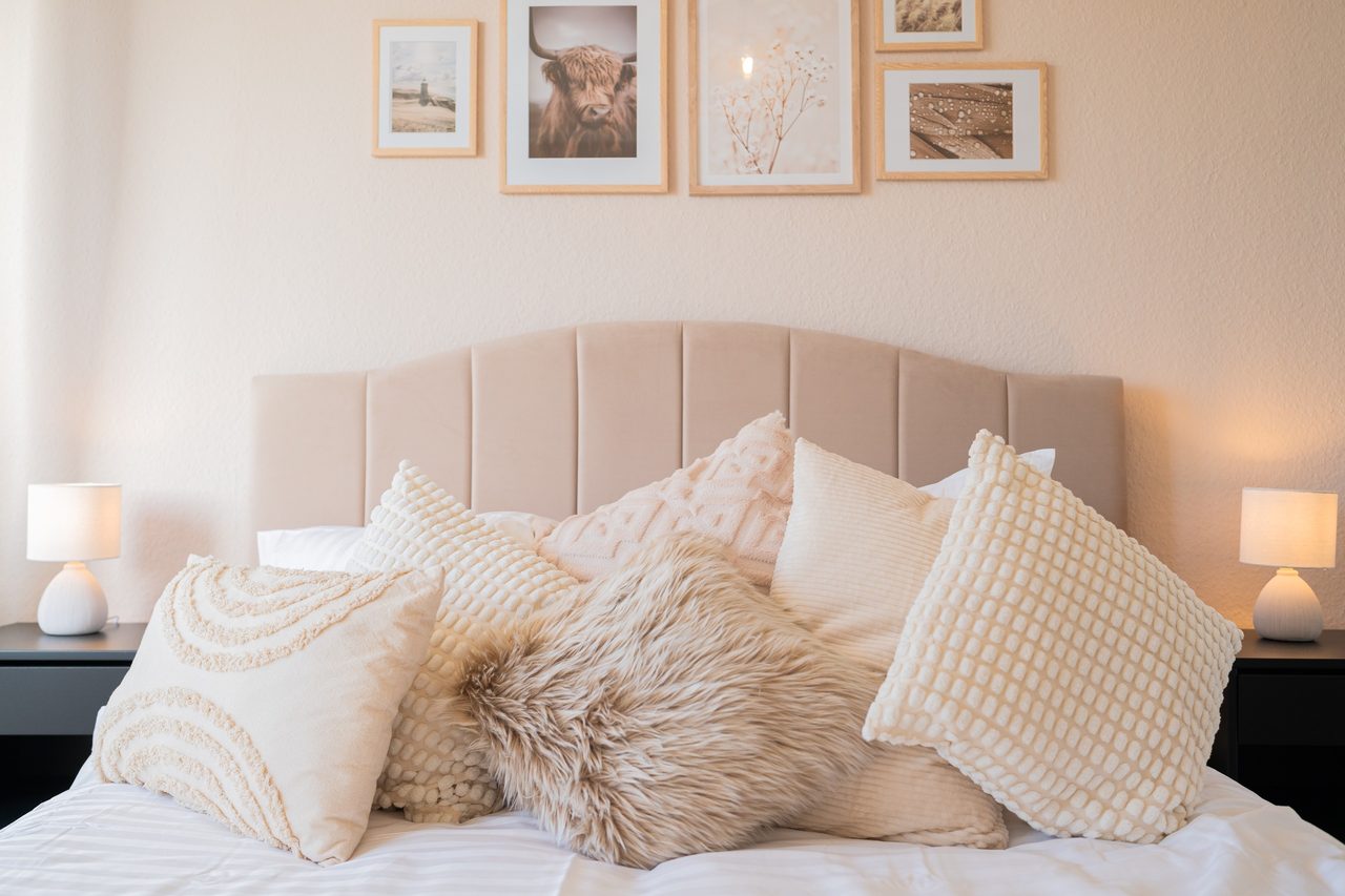 Bett mit beigem Polsterkopfteil und vielen hellen, flauschigen Kissen, links und rechts leuchten zwei kleine Nachttischlampen, darüber Bilder mit einem Hochlandrind.