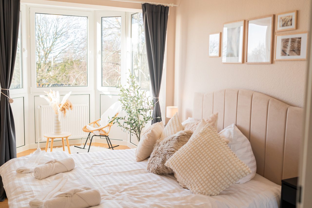 Helles Schlafzimmer mit Polsterbett, hellen Kissen und gerollten Handtüchern, links der Erker mit Fenstern, Rattansessel und kahlen Bäumen davor.