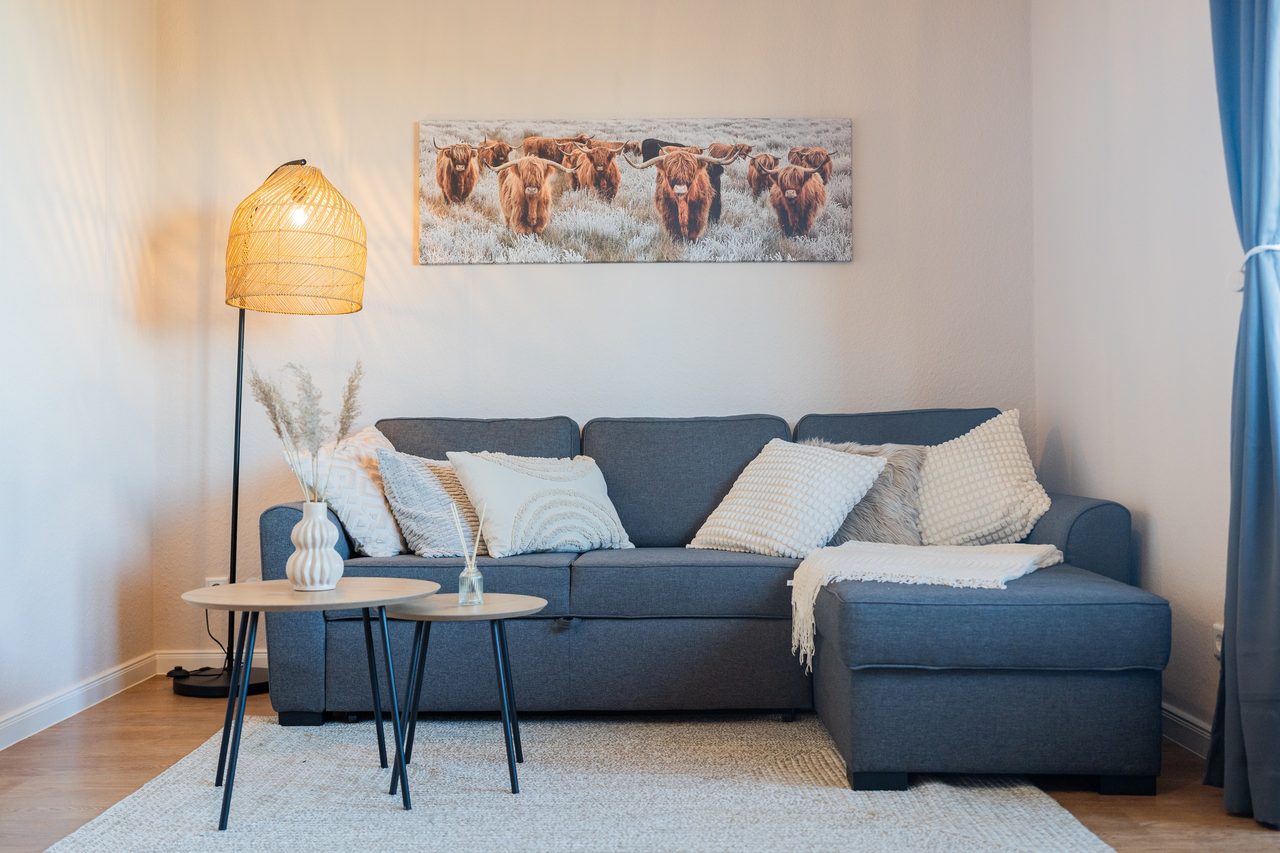 Gemütliche Sofaecke in Blau mit hellen Kissen, Rattan-Stehlampe und einem großen Wandbild mit Highland-Rindern, davor zwei runde Beistelltische.