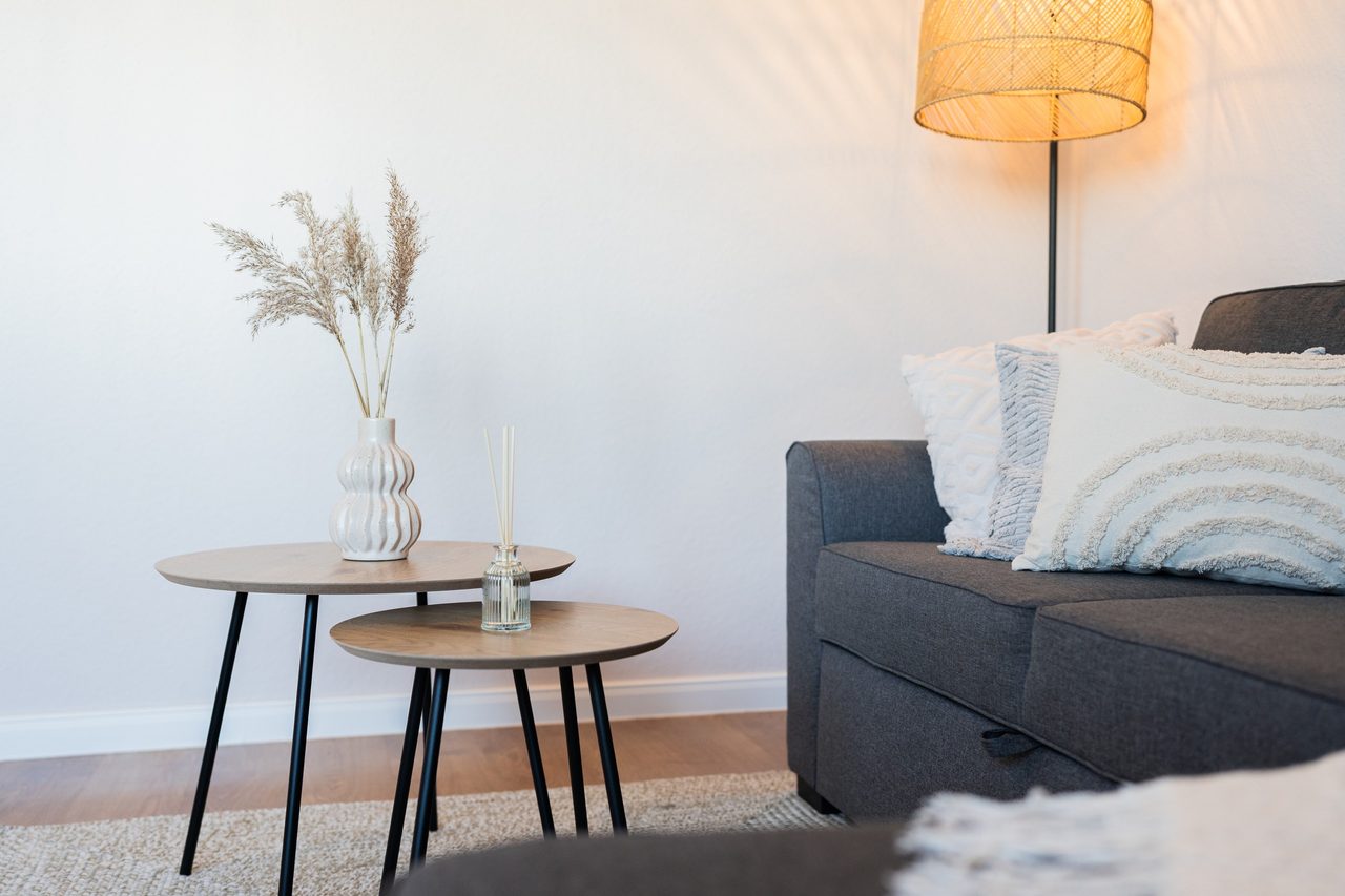 Helles Wohnzimmer in Küstenoptik: graues Sofa mit hellen, getufteten Kissen, zwei runde Holz-Beistelltische mit einer Vase Pampasgras und eine warm leuchtende Rattan-Stehlampe.