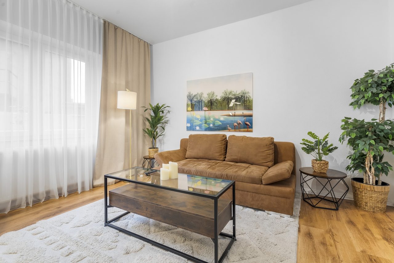 Wohnzimmer mit braunem Sofa, Glascouchtisch und großen Grünpflanzen