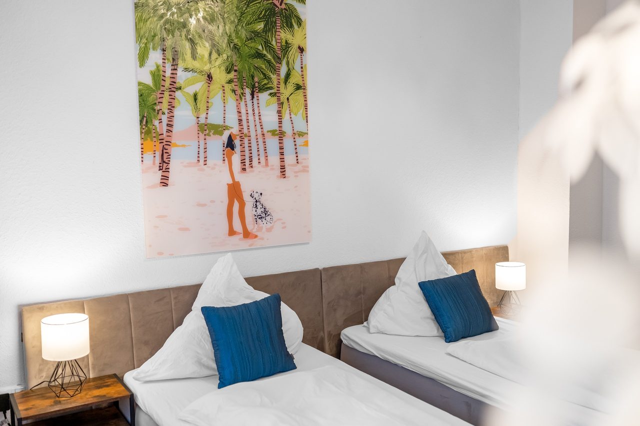 Schlafzimmer mit zwei Betten, blauen Kissen und Palmenbild