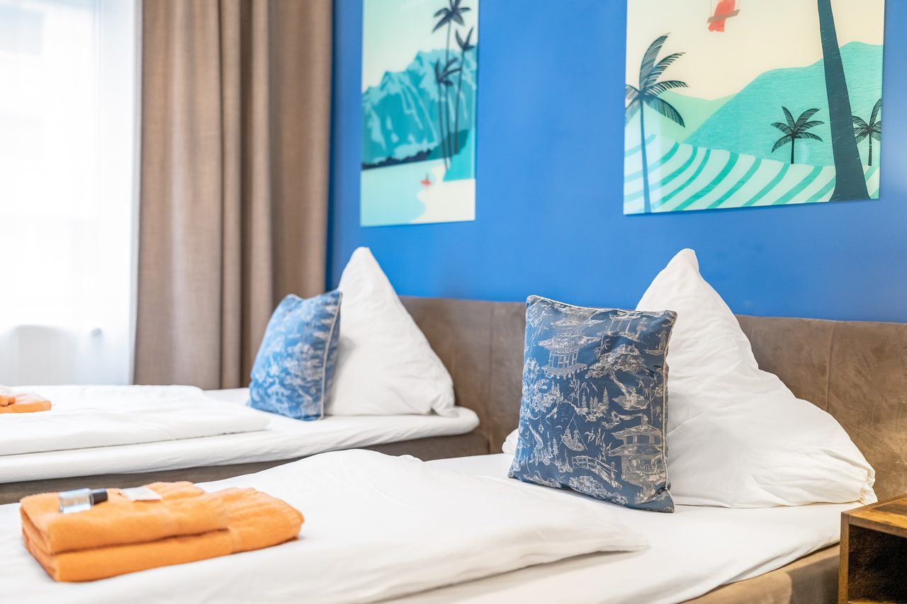 Schlafzimmer mit blauer Wand, zwei Betten und orangen Handtüchern