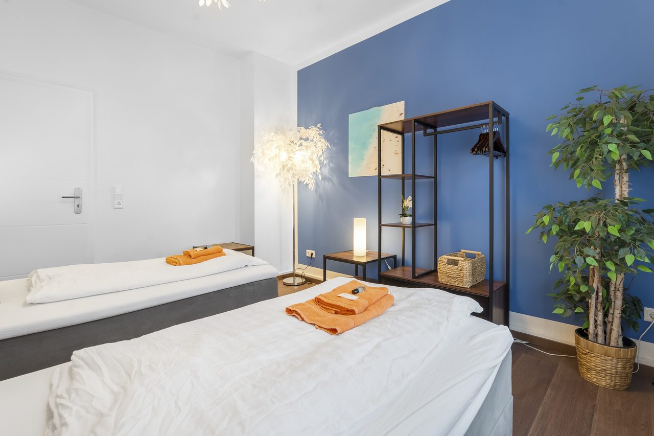 Schlafzimmer mit Einzelbetten, orangen Handtüchern und Grünpflanze