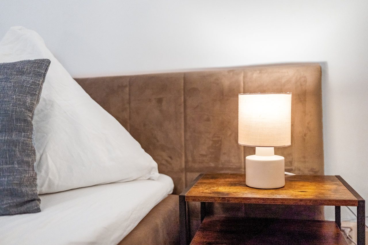 Nachttisch mit weißer Lampe neben Bett mit Polsterkopfteil