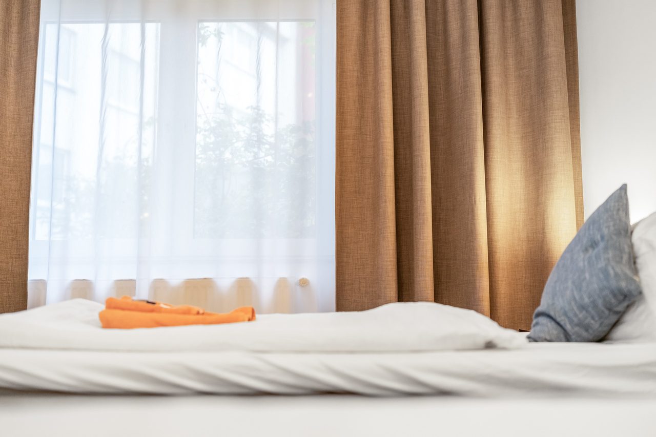 Bett mit weißer Bettwäsche, orangen Handtüchern und Vorhängen