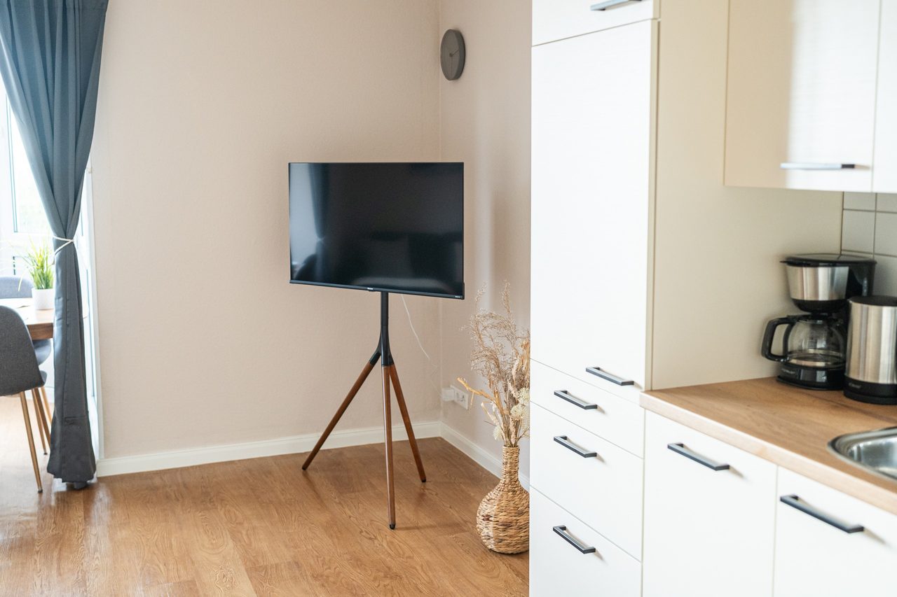 Wohnbereich mit Fernseher auf Dreibeinständer und Küchenzeile mit Kaffeemaschine