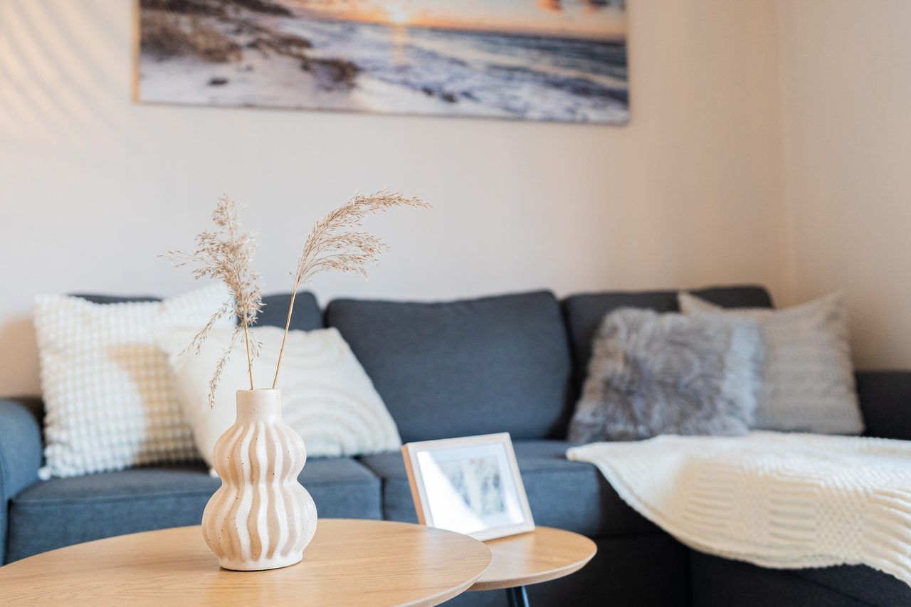 Blaues Sofa mit Kissen und Decke, davor Vase mit Trockengras