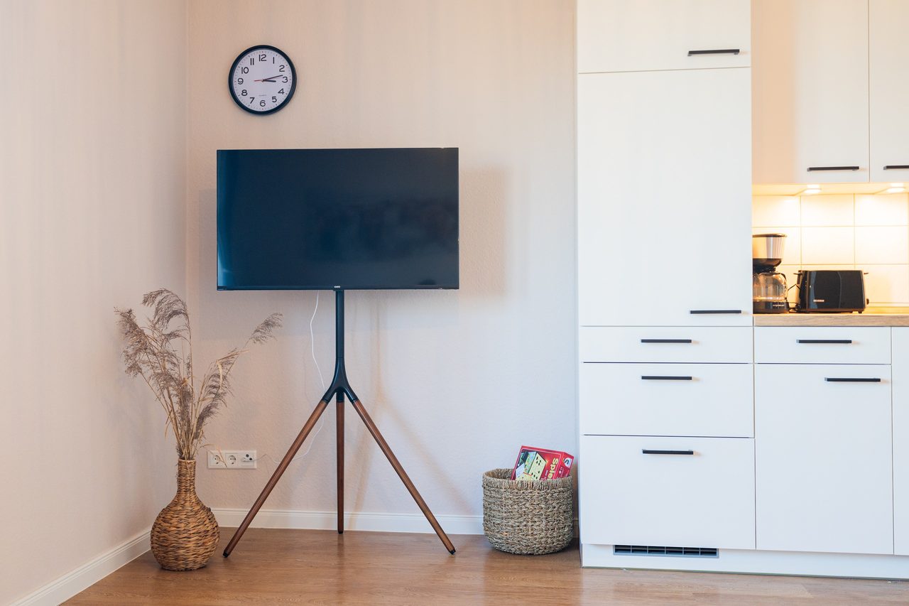 Wohnraum mit Fernseher auf Ständer, Wanduhr und weißer Küchenzeile