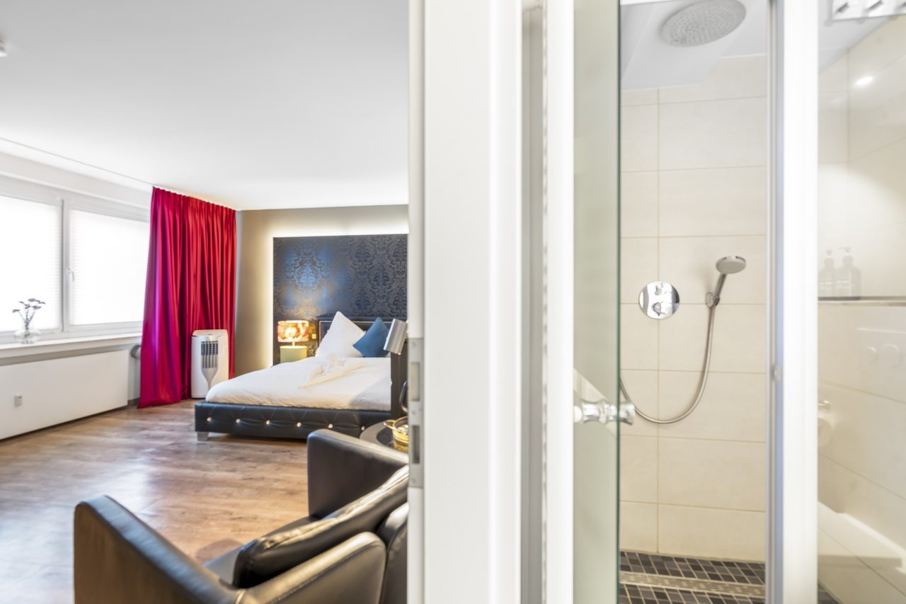 Blick durch die Glasdusche ins Schlafzimmer mit Doppelbett