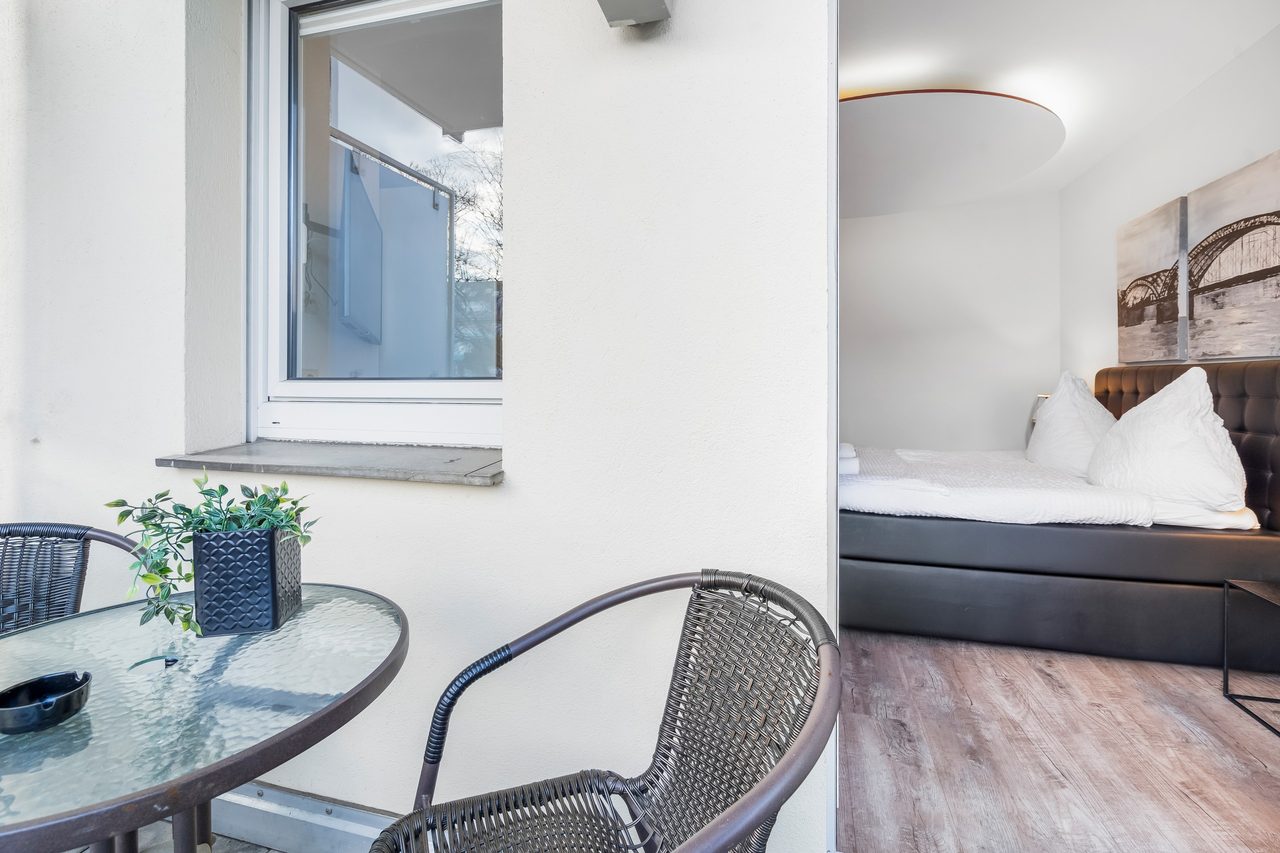 Balkon mit Glastisch und Rattanstuhl, daneben Blick auf ein Bett