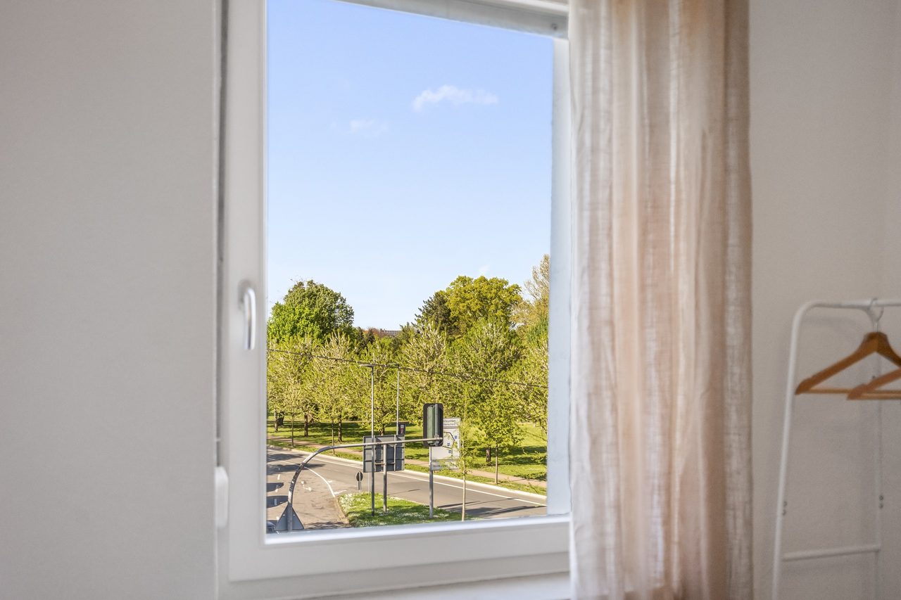 Fenster mit hellem Vorhang, Blick auf Bäume und Kleiderständer mit Bügel