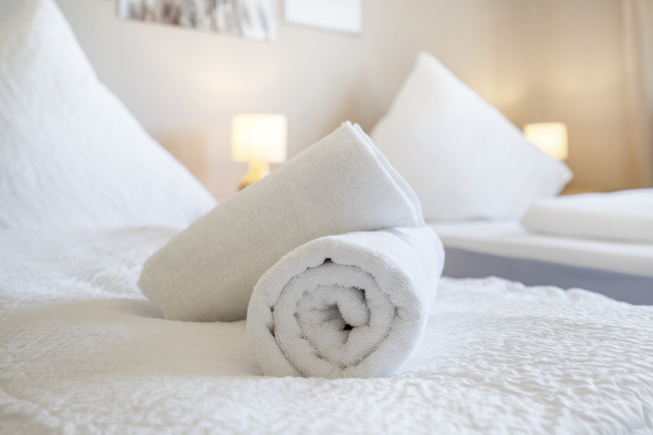 Gerolltes weißes Handtuch auf einem Bett mit Nachttischlampe im Hintergrund