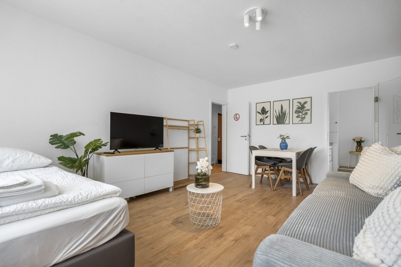Wohnraum mit Bett, Fernseher auf weißer Kommode und kleinem Esstisch