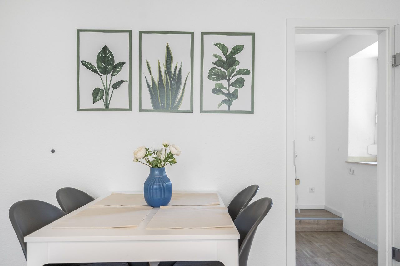 Esstisch mit grauen Stühlen, blauer Vase und drei Pflanzenbildern